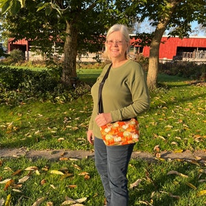 May include: A woman wearing a green jumper and blue jeans is standing in a grassy area with fallen leaves. She is carrying a small orange and white crossbody bag with a pumpkin pattern.