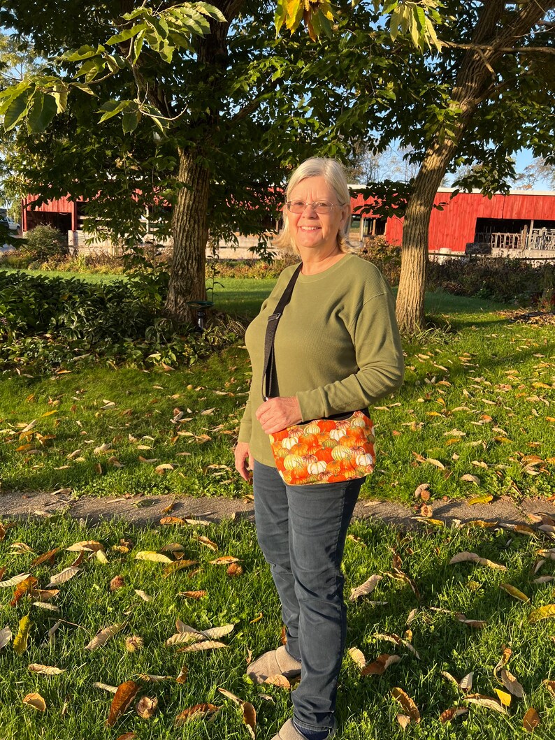 May include: A woman wearing a green jumper and blue jeans is standing in a grassy area with fallen leaves. She is carrying a small orange and white crossbody bag with a pumpkin pattern.