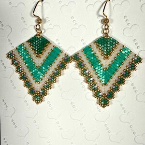 May include: A pair of handmade beaded earrings. Each earring features a geometric design with a teal, white, and gold color scheme. The earrings are square-shaped with a gold-colored hook.