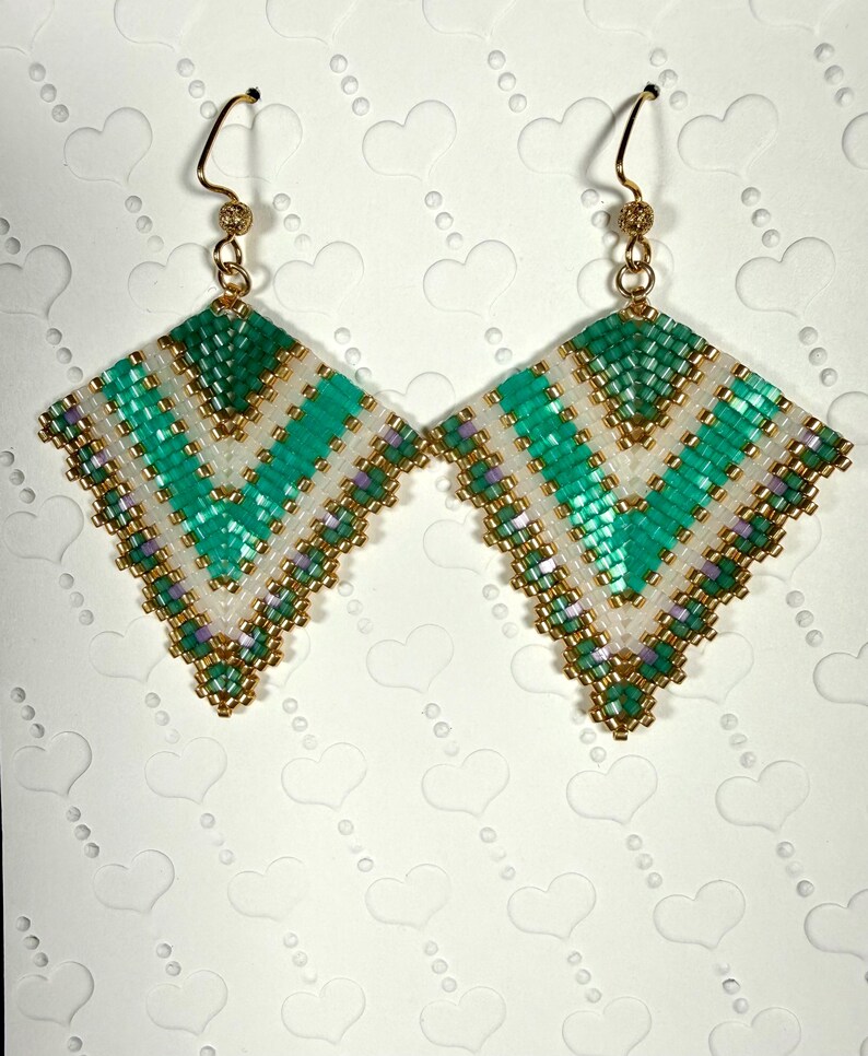 May include: A pair of handmade beaded earrings. Each earring features a geometric design with a teal, white, and gold color scheme. The earrings are square-shaped with a gold-colored hook.