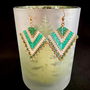 May include: A pair of handmade beaded earrings with a geometric design. The earrings feature a diamond shape with alternating rows of white, teal, and gold beads. The earrings are suspended from gold-colored ear wires.