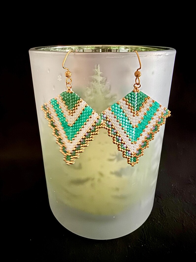 May include: A pair of handmade beaded earrings with a geometric design. The earrings feature a diamond shape with alternating rows of white, teal, and gold beads. The earrings are suspended from gold-colored ear wires.