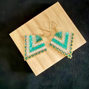 May include: A pair of handmade beaded earrings with a geometric design. The earrings feature a triangular shape with a pattern of green, white, and gold beads. The earrings are displayed on a wooden block against a black background.