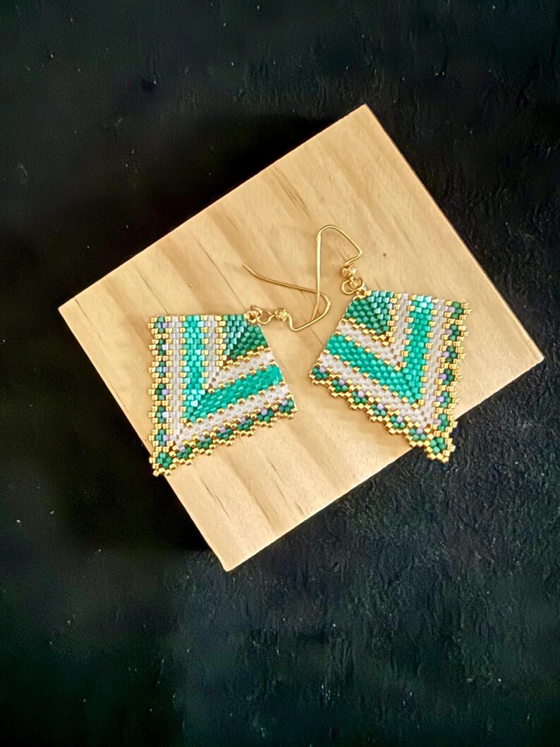 May include: A pair of handmade beaded earrings with a geometric design. The earrings feature a triangular shape with a pattern of green, white, and gold beads. The earrings are displayed on a wooden block against a black background.