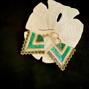 May include: A pair of handmade beaded earrings with a geometric design. The earrings feature a chevron pattern in shades of green, white, and gold. The earrings are square-shaped and have gold-colored ear wires. The earrings are displayed on a white, textured surface.
