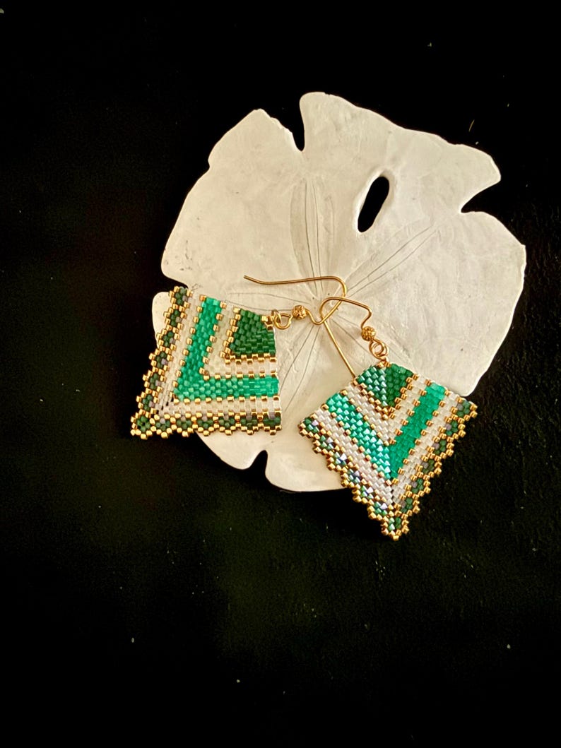May include: A pair of handmade beaded earrings with a geometric design. The earrings feature a chevron pattern in shades of green, white, and gold. The earrings are square-shaped and have gold-colored ear wires. The earrings are displayed on a white, textured surface.