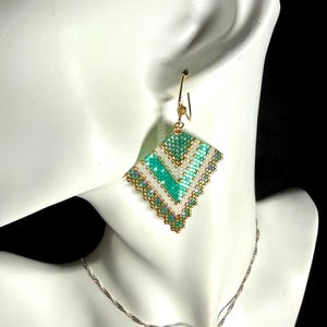 May include: A handmade, beaded earring with a geometric design. The earring features a diamond shape with a teal, white, and gold color scheme. The earring is suspended from a gold-colored hook. A silver necklace is also visible.