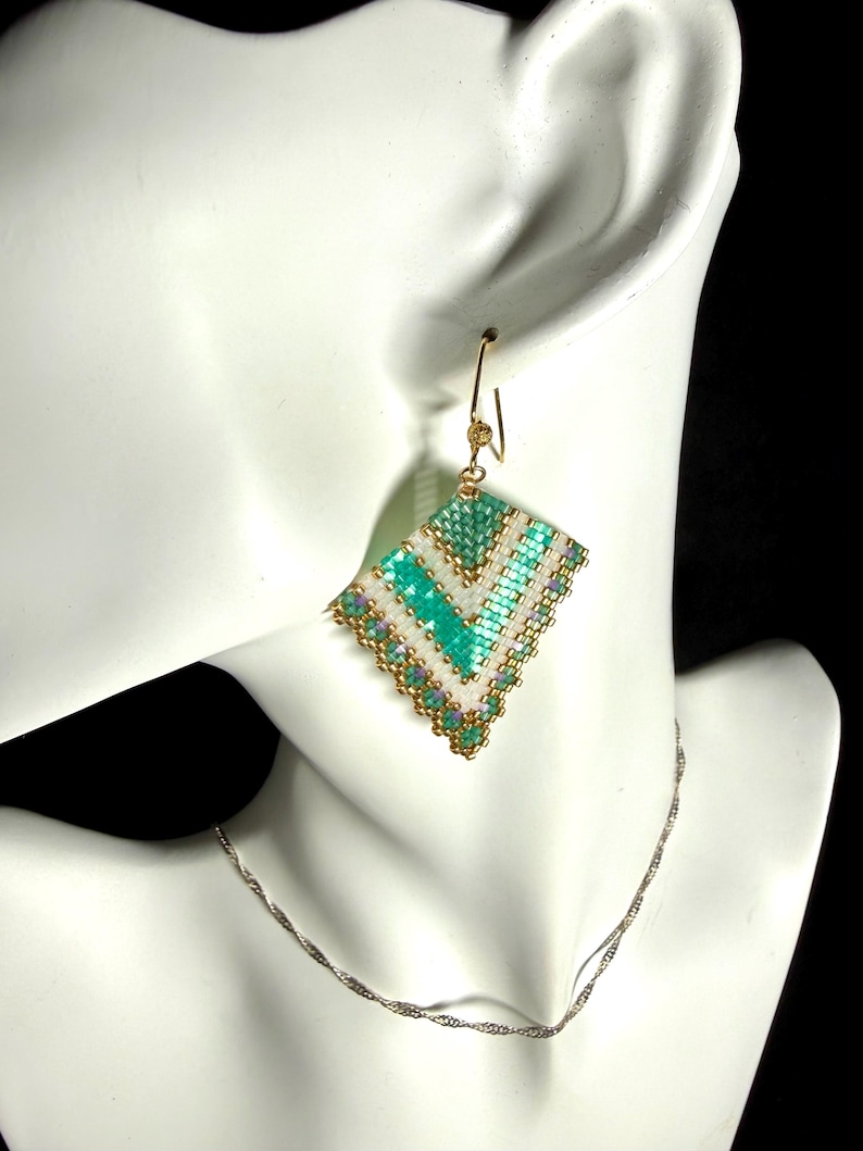 May include: A handmade, beaded earring with a geometric design. The earring features a diamond shape with a teal, white, and gold color scheme. The earring is suspended from a gold-colored hook. A silver necklace is also visible.