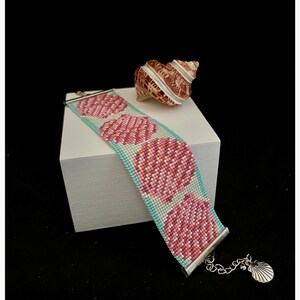 May include: A beaded bracelet with a pink and white seashell design on a turquoise background. The bracelet has a silver clasp and a small silver seashell charm.