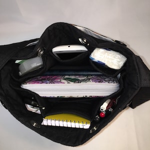 May include: A black canvas tote bag with multiple pockets and compartments. The bag is open and filled with various items, including a phone, a notebook, pens, and a small makeup bag.