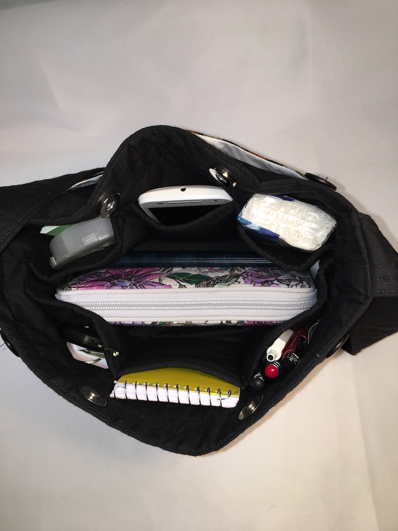 May include: A black canvas tote bag with multiple pockets and compartments. The bag is open and filled with various items, including a phone, a notebook, pens, and a small makeup bag.