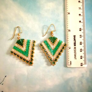 May include: A pair of handmade beaded earrings with a geometric design. The earrings feature a diamond shape with green, white, and gold beads. The earrings are approximately 1.5 inches in length and have gold-colored ear wires.
