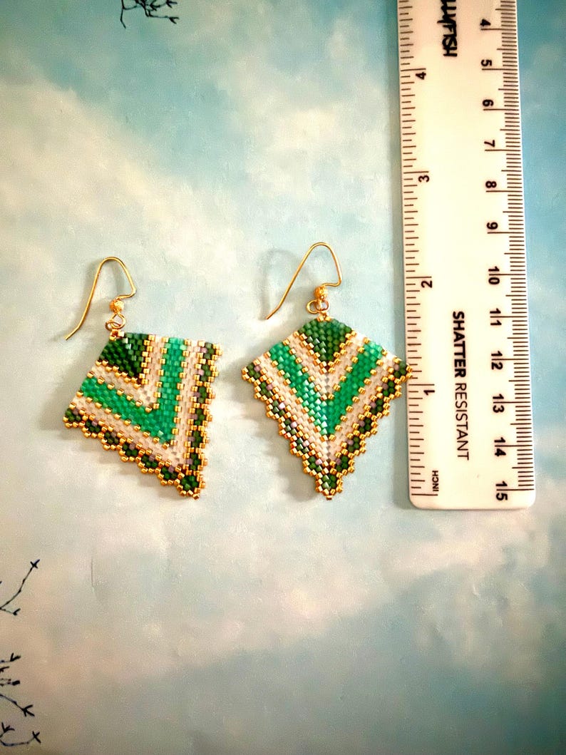 May include: A pair of handmade beaded earrings with a geometric design. The earrings feature a diamond shape with green, white, and gold beads. The earrings are approximately 1.5 inches in length and have gold-colored ear wires.