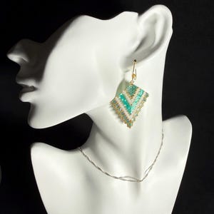 May include: A pair of beaded earrings in a diamond shape, featuring turquoise, white, and gold beads. The earrings are displayed on a white mannequin head, with a silver necklace. The background is black.