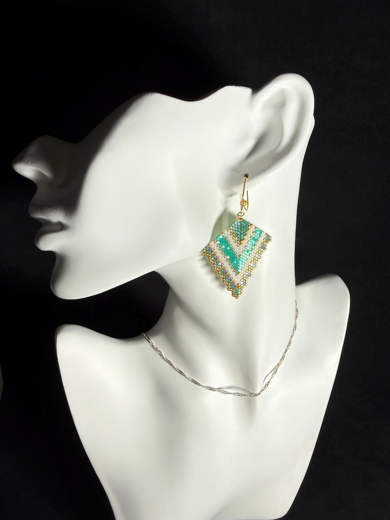 May include: A pair of beaded earrings in a diamond shape, featuring turquoise, white, and gold beads. The earrings are displayed on a white mannequin head, with a silver necklace. The background is black.