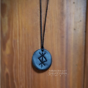 May include: A black pendant necklace featuring a circular, dark gray stone with an etched rune symbol. The pendant hangs from a thin, black cord. The background is a wooden surface. The text "Solitaire Bon Lavi BonLaviCreations" is visible.