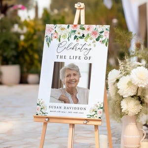 May include: A wooden easel displays a sign with the text "Celebrating the Life of" above a photo of a woman. Floral arrangements in pink and white decorate the top and bottom of the sign. The sign includes the name "Susan Davidson" and dates.