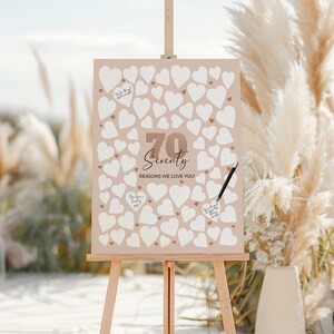 Può includere: Un cavalletto in legno espone un cartello con sfondo beige e cuori bianchi. Il cartello recita "70 Seventy Reasons We Love You!" in una scrittura elegante. Una penna nera è appoggiata sul cartello, pronta per gli ospiti per scrivere messaggi.