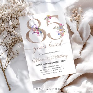Könnte beinhalten: Eine weiße Einladungskarte mit dem Text "85 Jahre geliebt" und "Maria's 85. Geburtstag". Die Zahlen "85" sind goldfarben und mit bunten Blumen verziert. Die Karte ist für eine Geburtstagsfeier.