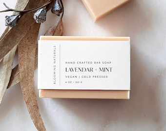 May include: A bar of hand-crafted lavender and mint soap. The soap is a light peach color and is wrapped in white paper with black text that reads "BLOOMING NATURALS", "HAND CRAFTED BAR SOAP", "LAVENDAR + MINT", "VEGAN | COLD PRESSED", and "4 OZ - 120 G".