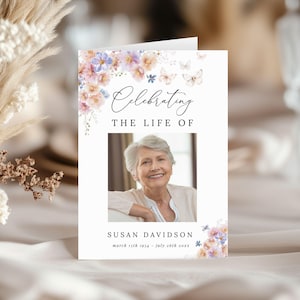 May include: A memorial card with the text "Celebrating The Life Of" above a photo of a smiling woman. The card is decorated with floral and butterfly illustrations in shades of pink, purple, and blue. The name "Susan Davidson" and dates are below the photo.