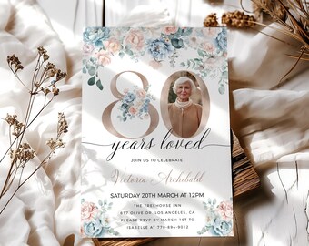 May include: A floral-themed 80th birthday invitation with a photo of a woman in a brown jacket. The invitation is decorated with blue and pink flowers and greenery. The text on the invitation reads "80 years loved" and "Join us to celebrate Victoria Archibald". The invitation also includes the date, time, and location of the event.