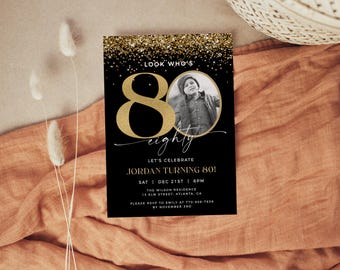 May include: Black and gold 80th birthday invitation. The invitation features a gold glitter top border, a large gold "80", and a black and white photo. Text includes "Look Who's", "Let's Celebrate", and event details.