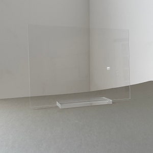 May include: A clear acrylic picture frame with a rectangular base. The frame is standing on a grey surface.