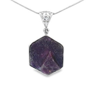 Puede incluir: Un collar de plata con un colgante hexagonal. El colgante presenta una gema púrpura rugosa engastada en un marco de plata, suspendida de una fina cadena de plata. La gema tiene forma de pirámide en el centro.
