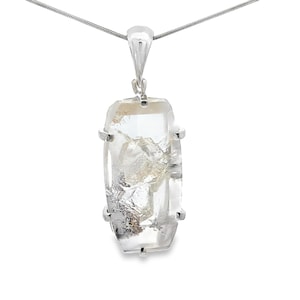 Puede incluir: Un colgante de cuarzo transparente con engaste y cadena plateados. El cuarzo es rectangular con inclusiones naturales y facetas. El colgante cuelga de una delicada cadena plateada, creando un diseño minimalista y elegante.