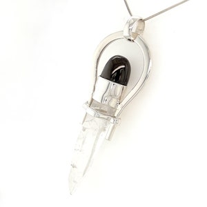May include: A silver pendant with a clear quartz crystal and a black gemstone. The pendant is designed in a unique shape with a loop at the top for attaching a chain.