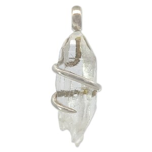 Puede incluir: Un colgante de cristal de cuarzo transparente envuelto en alambre de plata. El cristal es largo y estrecho con una punta puntiaguda. El alambre está envuelto alrededor del cristal en espiral.