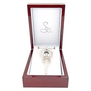 May include: A silver pendant with a clear quartz crystal point and a black stone at the top, displayed in a red wooden box with a white interior and the letter "S" in a cursive font.