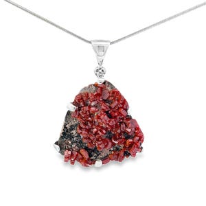 Może przedstawiać: Srebrny naszyjnik z wisiorkiem z surowym, nieregularnym kamieniem. Kamień jest ciemnoszary z skupiskami żywych, głęboko czerwonych kryształów. Wisiorek zawieszony jest na delikatnym srebrnym łańcuszku.