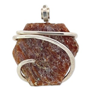 Puede incluir: Un colgante de piedra preciosa de color marrón rojizo en bruto envuelto en alambre de plata. El colgante tiene forma hexagonal y una textura rugosa y natural.