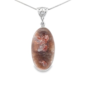 Puede incluir: Un collar con colgante ovalado que presenta una piedra marrón translúcida con inclusiones de color marrón rojizo. La piedra está engastada en un marco de metal plateado con una pequeña piedra clara en la argolla, suspendido de una cadena fina.