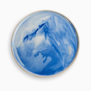 Pode incluir: Prato redondo de cerâmica com um design marmorizado azul e branco. O prato tem uma borda bege clara. O padrão azul assemelha-se a água corrente ou a um efeito de aguarela. Uma peça decorativa para servir alimentos, cerca de 25 cm de diâmetro.