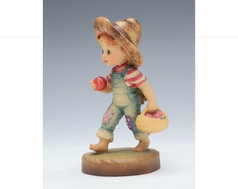 May include: A wooden figurine of a child wearing a straw hat, overalls, and a red and white striped shirt. The child is holding a red apple in one hand and a basket of apples in the other. The figurine is standing on a wooden base.