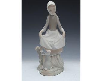 May include: A porcelain figurine of a girl in a white dress and brown waistcoat, wearing a white headscarf. She is holding her skirt and looking down at two rabbits, one white and one brown, standing on a base.