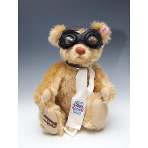 May include: A tan teddy bear wearing a black leather aviator hat and goggles, a white scarf, and a patch on its foot that says "Wright Aircraft 100th Anniversary 1903-2003".