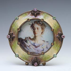 May include: A round, ornate picture frame with a green and gold enamel finish. The frame is decorated with rhinestones and features a portrait of a woman in a white dress.