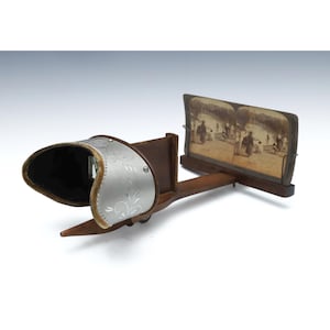 May include: Antique stereoscope viewer with a silver-colored eye shield featuring an engraved floral design. The viewer has a wooden frame and holds a vintage stereoscopic image of people. The viewer is designed to provide a 3D effect.