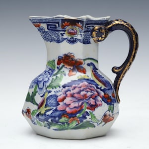 May include: A ceramic pitcher featuring a white base with a multi-colored floral and bird pattern in shades of blue, pink, red, and green. The handle is dark blue with gold accents and a serpent-like head detail at the top attachment point.