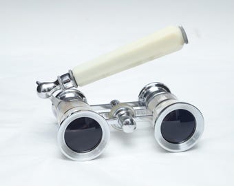 May include: Vintage opera glasses featuring a polished chrome metal frame with two circular black lenses and a central focusing knob. An off-white, hexagonal-shaped handle is attached to the side by a hinged joint, allowing for adjustable positioning.