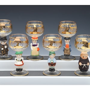May include: Set of seven clear glass wine glasses with gold accents and hand-painted figurines. The figurines are dressed in traditional European attire.