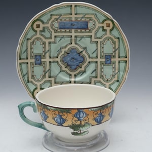 May include: A white teacup and saucer with a green and blue garden design. The saucer features a geometric pattern of a garden with a central fountain. The teacup has a yellow and blue border with a garden scene.