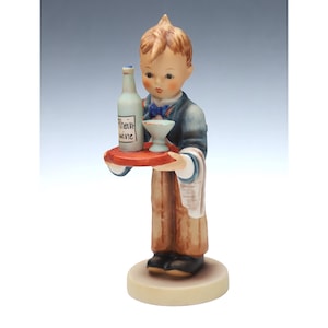 Puede incluir: Una figurita de porcelana de un joven camarero que sostiene una bandeja con una botella de vino y una copa. La botella está etiquetada con "Rhein wine".
