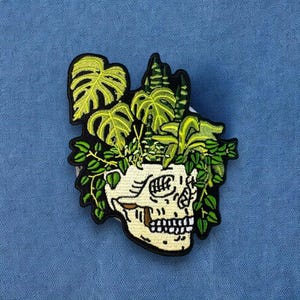 May include: A white skull with a green and yellow plant growing out of the top of its head. The skull is embroidered on a fabric patch.