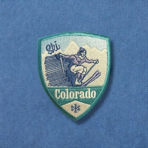 May include: A green and white embroidered patch with a skier in a blue and purple outfit on a mountain with the text "Ski Colorado" and a snowflake.