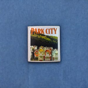 May include: A white patch with a colorful image of houses and a mountain range. The text "PARK CITY" is in orange letters at the top of the patch. The text "Utah" is in smaller black letters below the city name.